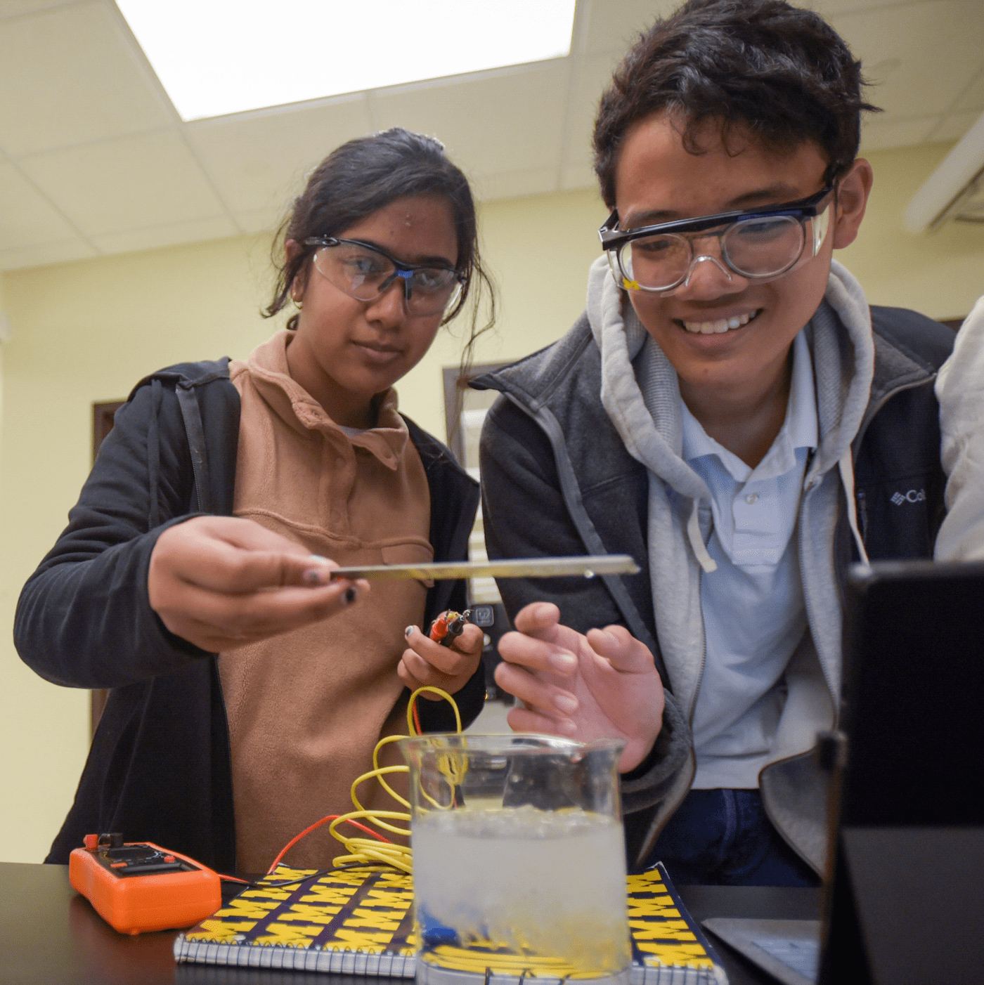 homepage3-min Two students in safety goggles looking over beaker conducting experiment